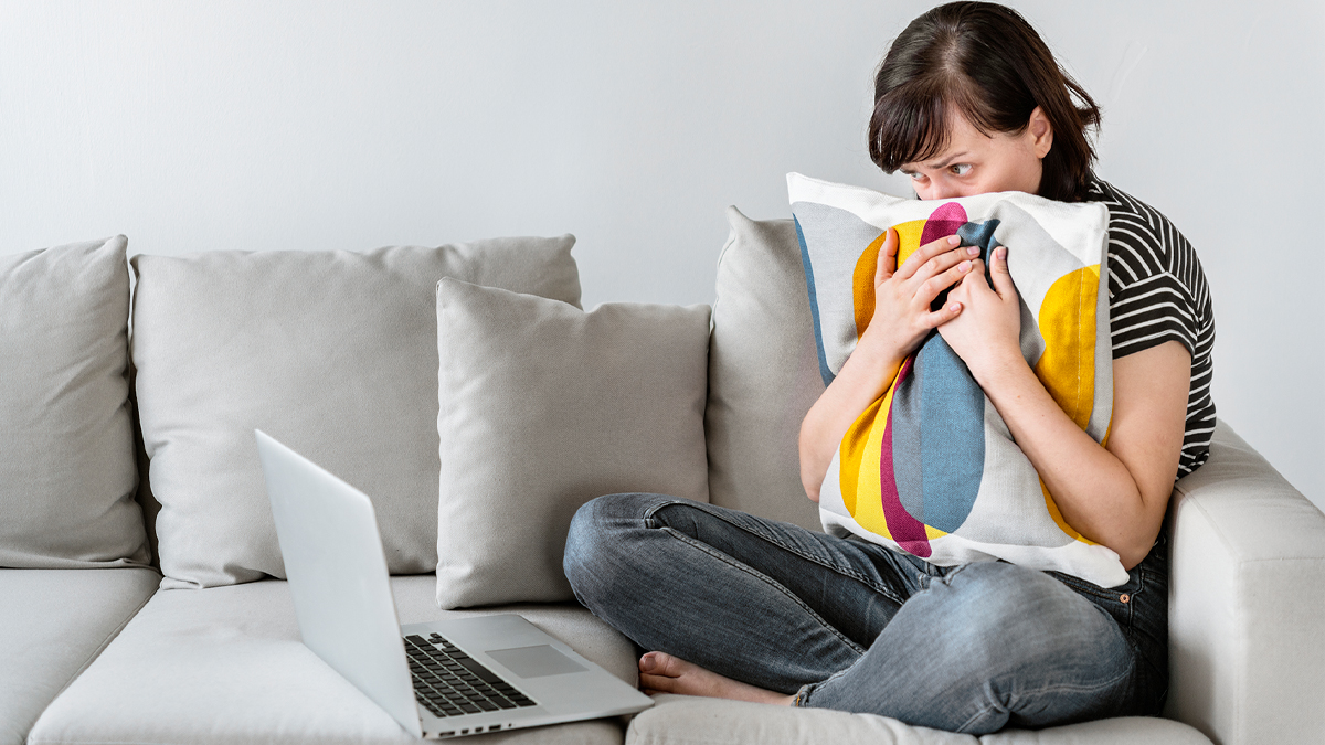 Woman on couch, hiding behind pillow and afraid of what she's reading on computer screen